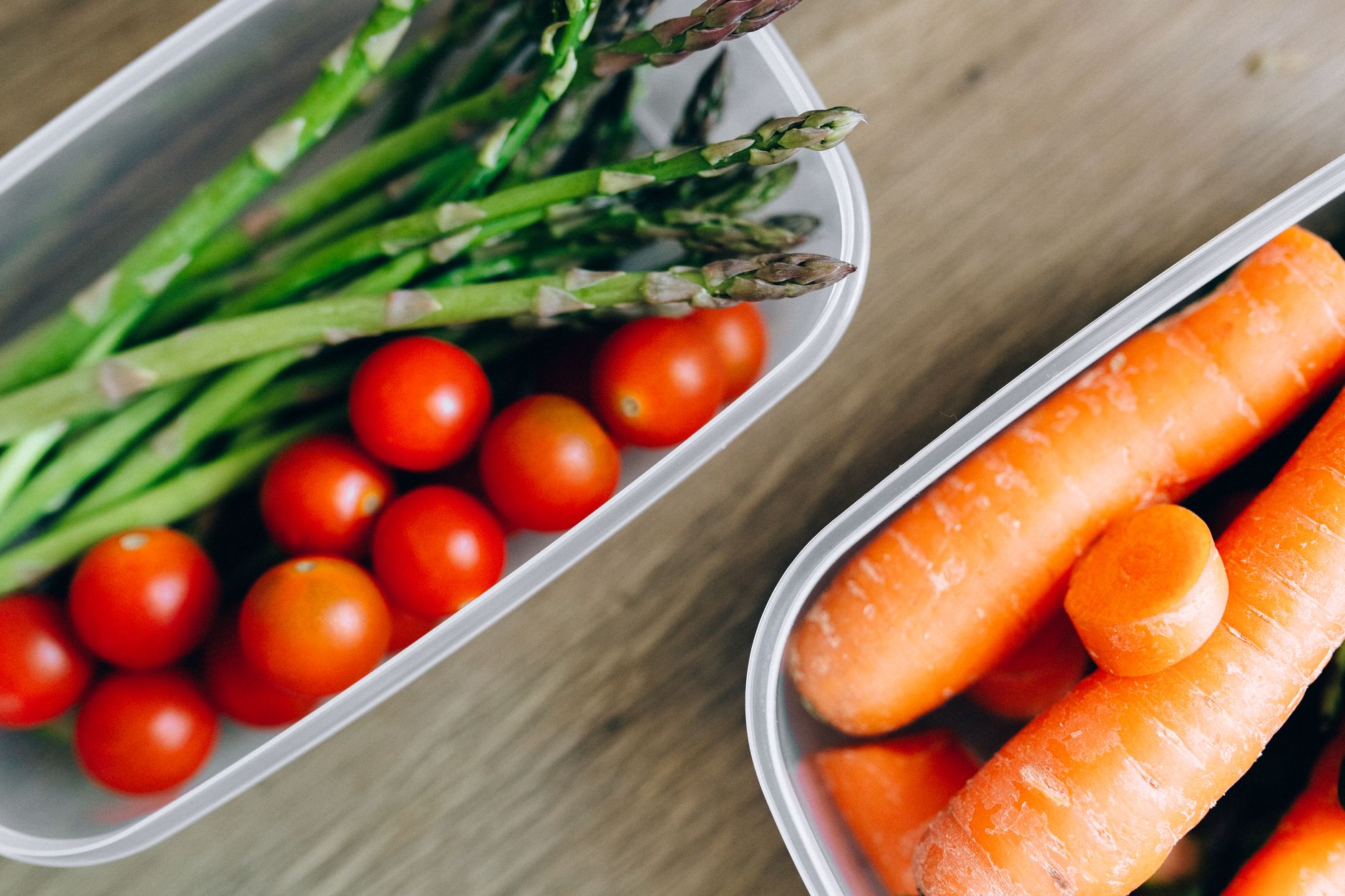 Close-up of fresh asparagus, cherry tomatoes, and carrots in plastic containers on a wooden table.