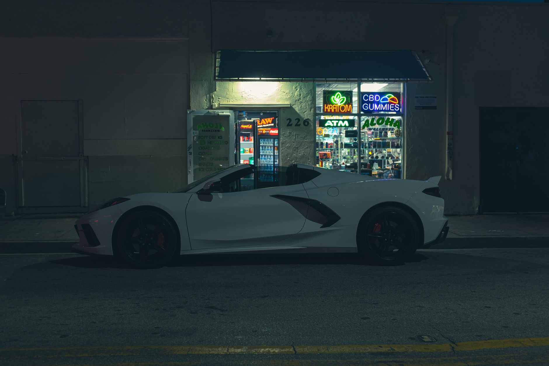 White sports car parked at night outside an illuminated CBD gummies store with neon signs.