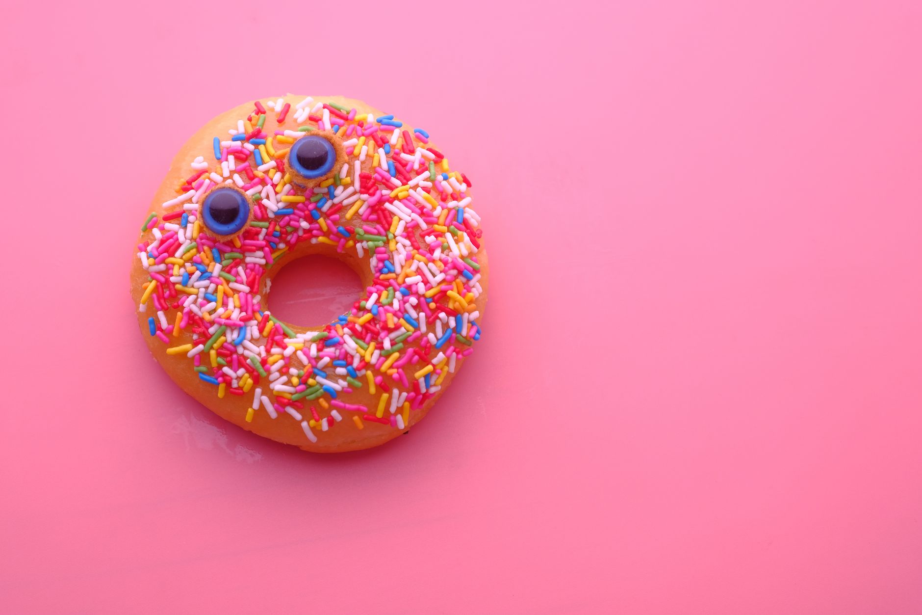 Vibrant donut with sprinkles and candy eyes on a pink backdrop, perfect for food enthusiasts.