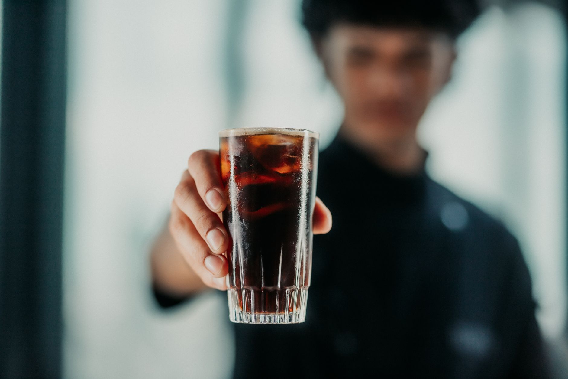 Close-up of a person offering a glass of dark beverage with ice, indoors.